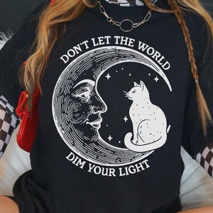 A person wearing a black t-shirt with a graphic design of a cat on a crescent moon and the text "Don't Let the World Dim Your Light".