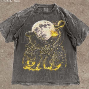 Może przedstawiać: Ciemnoszara koszulka z grafiką przedstawiającą trzy kolczaste dinozaury na tle pełni księżyca, z żółtymi i pomarańczowymi akcentami. Na koszulce nadrukowano tekst "THE SILLY RACCOON CO" i "DARK GREY".