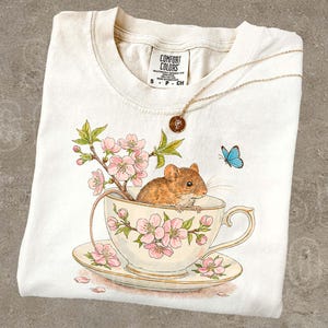 May include: Cream-colored t-shirt featuring a whimsical illustration of a mouse in a teacup with cherry blossoms and a blue butterfly. A gold necklace with a pendant rests on the shirt. The text "THE SILLY RACCOON CO" is at the top left.