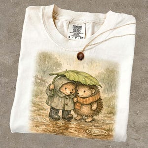 A white t-shirt with a printed illustration of two hedgehog characters standing together under an umbrella, with one hedgehog wearing a raincoat.