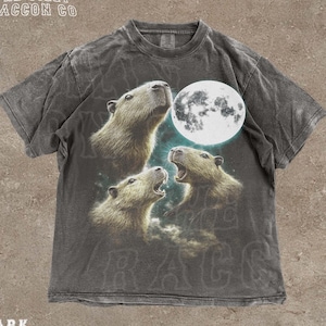 May include: Dark grey t-shirt featuring a graphic of three capybaras looking up at a full moon. The shirt has the text "THE SILLY RACCOON CO" at the top and "DARK GREY" at the bottom. The shirt has a vintage, distressed look.