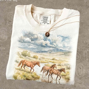 May include: A cream-colored t-shirt with a watercolor painting of horses in a river, under a cloudy sky. The shirt has a round neckline and short sleeves. A gold necklace with a round pendant rests on the shirt. The text "THE SILLY RACCOON CO" is at the top left.
