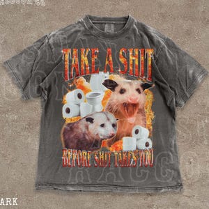 A t-shirt with a graphic design featuring an opossum surrounded by toilet paper rolls, with the text "TAKE A SHIT BEFORE SHIT TAKES YOU" prominently displayed.
