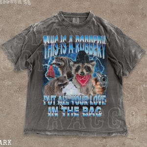 May include: Dark grey t-shirt featuring a graphic of raccoons, one in a cowboy hat and red bandana, holding a gun. Text on the shirt says "THIS IS A ROBBERY" and "PUT ALL YOUR LOVE IN THE BAG". The shirt has a distressed, vintage appearance.