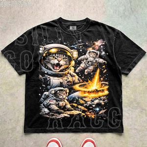 May include: Black t-shirt featuring a graphic of cats in astronaut suits exploring space. The design includes a large cat in a spacesuit, two smaller cats, and a cosmic explosion. The text "THE SILLY RACCOON CO" is at the top.