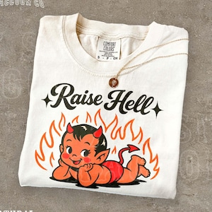 May include: Cream-colored t-shirt with the text "Raise Hell" and a cartoon devil graphic. The devil is red and orange, surrounded by flames. A gold necklace with a pendant rests on the shirt.