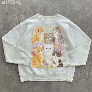 May include: White sweatshirt featuring a graphic of six kittens in shades of orange, grey, and white, set amongst flowers. The text "THE SILLY RACCOON CO" is visible, with "WHITE" at the bottom. The garment has a crew neck and long sleeves.