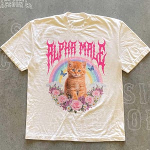 Może przedstawiać: Kremowa koszulka z grafiką pomarańczowego kociaka z niebieskimi oczami, tęczą, motylami i kwiatami. Napis "ALPHA MALE" jest w kolorze różowym, gotycką czcionką. Koszulka pochodzi od "THE SILLY RACCOON CO."