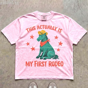 A pink t-shirt with a graphic of a green horse and the text "This actually is my first rodeo" printed on it.