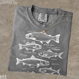 May include: Dark grey t-shirt with a white graphic of various fish species, including rainbow trout, largemouth bass, and salmon. The shirt has a crew neck and a small tag that says "S P CH". The text "DARK GREY" is at the bottom.