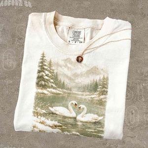 A white t-shirt with a printed image of two swans swimming in a lake, surrounded by snow-covered trees.