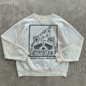 May include: Ash-colored sweatshirt with a graphic of a raccoon wearing a party hat. The raccoon is inside a black rectangular frame. The text "Grilled cheese destroyer" is below the raccoon. The text "THE SILLY RACCOON CO" is at the top.
