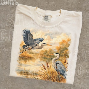 Könnte beinhalten: Cremefarbenes T-Shirt mit einer detaillierten Illustration von zwei Reihern in der Nähe eines Sees. Ein Reiher ist im Flug, die Flügel ausgebreitet, der andere steht zwischen Schilf. Das Shirt hat den Text "THE SILLY RACCOON CO" oben.