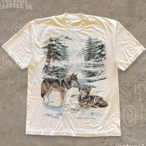 A white t-shirt with a printed design depicting two wolves in a snowy forest landscape.
