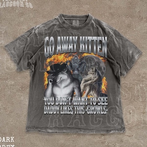 Puede incluir: Camiseta gris oscuro con un gráfico que presenta un lobo con sombrero, llamas y el texto "GO AWAY KITTEN" y "YOU DON'T WANT TO SEE DADDY LIKES THIS - GROWLS." La camiseta tiene un aspecto vintage y desgastado.
