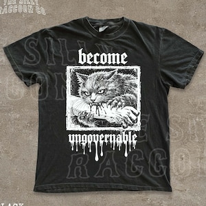 Puede incluir: Camiseta negra con un gráfico desgastado de un gato gruñón mordiendo algo, con las palabras "become ungovernable" en fuente gótica. La camiseta también tiene el texto "THE SILLY RACCOON CO" en la parte superior.