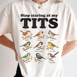 May include: Cream-colored t-shirt with the text "Stop staring at my TITS" in black. Below the text are illustrations of various birds, each labeled with its name. The shirt is a casual, unisex style.