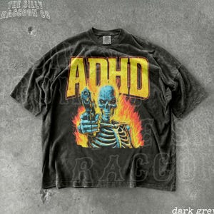 Könnte beinhalten: Dunkelgraues T-Shirt mit einem Used-Look-Grafikdesign, das ein Skelett mit einer Pistole, Flammen und dem Wort "ADHD" in Gelb zeigt. Das Shirt hat einen Vintage-Look und der Text "THE SILLY RACCOON CO" ist oben links aufgedruckt.