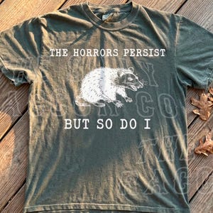 A t-shirt with a humorous text printed on it, lying on a wooden surface with some fallen leaves around it.
