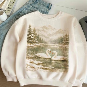 A white sweatshirt with a printed image of two swans swimming in a snowy lake, surrounded by trees and mountains.