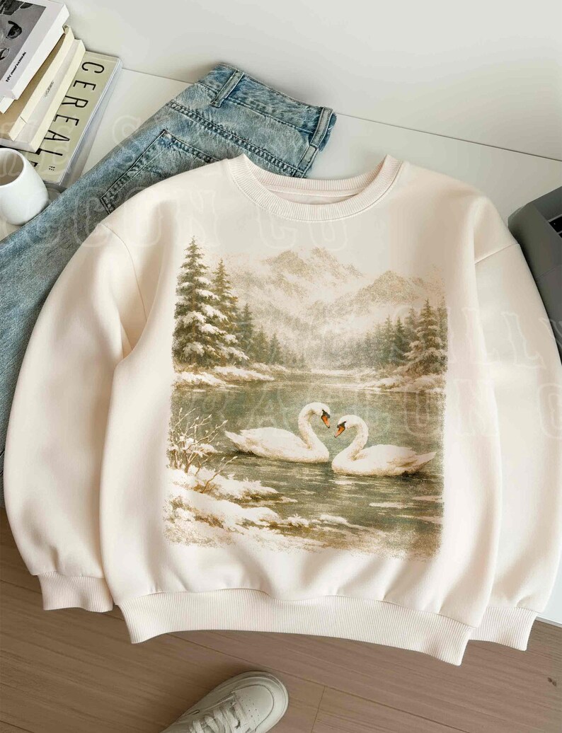 A white sweatshirt with a printed image of two swans swimming in a snowy lake, surrounded by trees and mountains.
