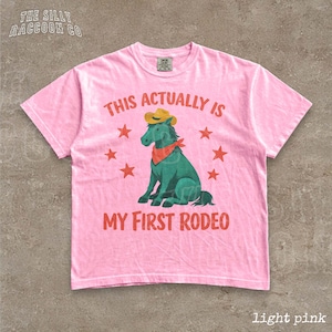 A pink t-shirt with a graphic design depicting a green horse wearing a cowboy hat and surrounded by red stars. The text on the t-shirt reads "This actually is my first rodeo".