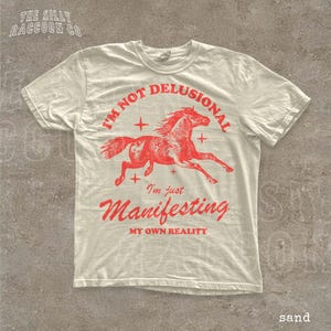 A white t-shirt with a red graphic design depicting a running horse and the text "I'm not delusional, I'm just Manifesting my own reality" printed on it.