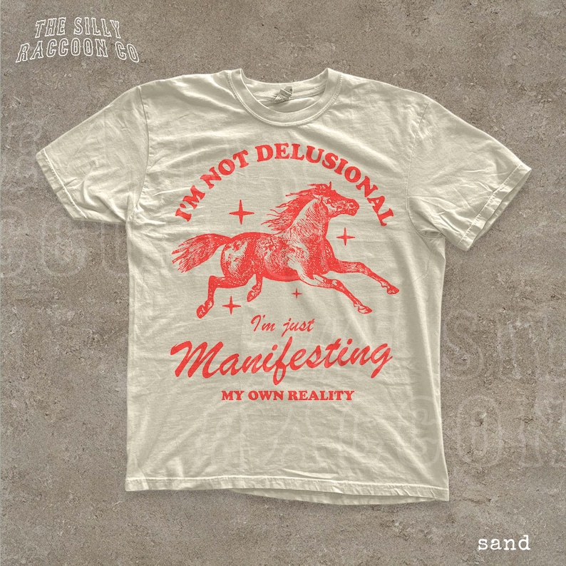 A white t-shirt with a red graphic design depicting a running horse and the text "I'm not delusional, I'm just Manifesting my own reality" printed on it.