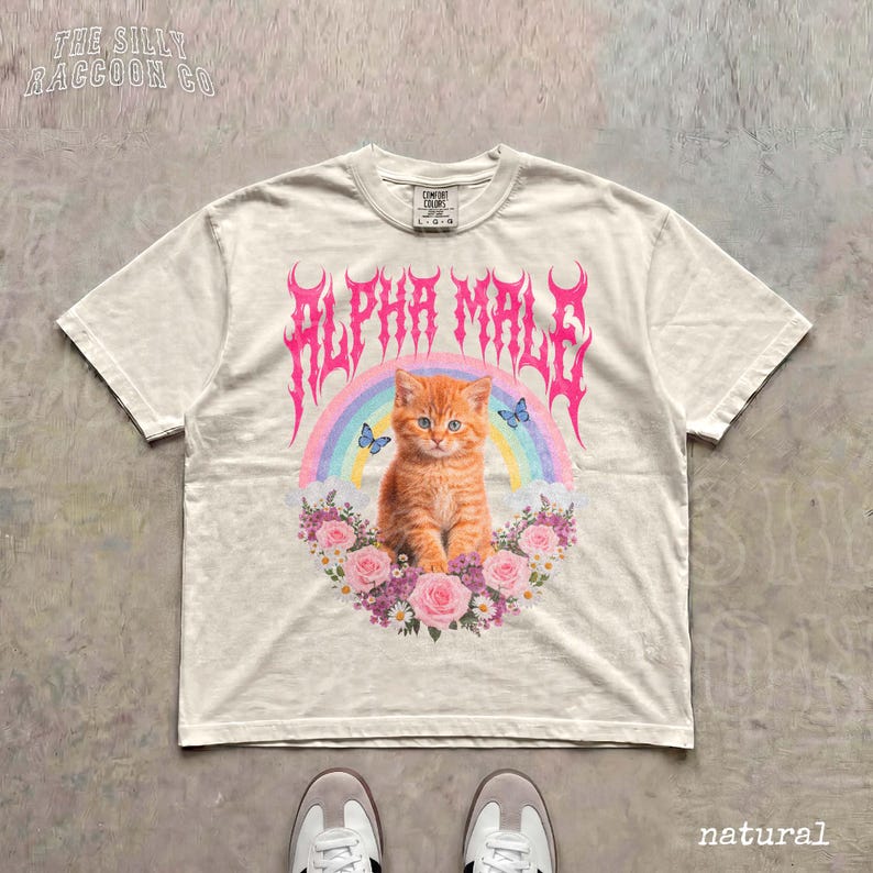 A white t-shirt with a graphic design featuring an orange cat surrounded by flowers and a rainbow, with the text "ALPHA MALE" prominently displayed.