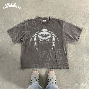 A pair of sneakers and a gray t-shirt with a spider design on it, laid out on a concrete floor.