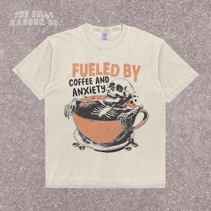 Funny coffee shirt for anxious introverts, featuring a skeleton relaxing in a giant coffee cup with the text “Fueled by Coffee and Anxiety.” Vintage grunge design with distressed print and muted tones. Ideal for women, men, and non-binary adults who love dark humor and caffeine culture. Great for Halloween, work-from-home vibes, mental health awareness, or gifting your favorite goth coffee lover.