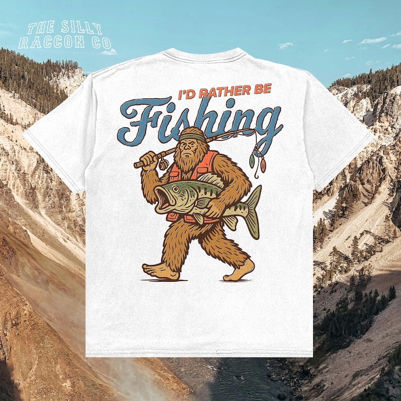 A white t-shirt with a graphic design depicting a large, furry creature holding a fish and the text "I'D Rather Be Fishing" against a mountainous background.