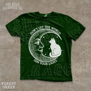 A green t-shirt with a white illustration of a cat sitting on a crescent moon and the text "Don't Let the World Dim Your Light" printed on it.
