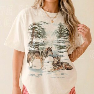 A woman wearing a white t-shirt with a graphic design of two wolves in a snowy forest.