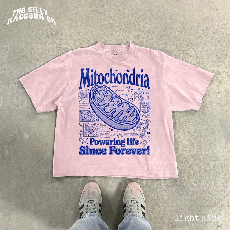 A pink t-shirt with a graphic design depicting Mitochondria, a cellular organelle, and the text "Powering life since forever!" printed on it. The t-shirt is laid out on a gray floor, and a pair of feet is visible in the bottom right corner of the image.