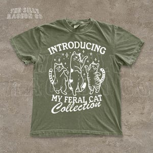 A green t-shirt with a graphic design featuring three cats and the text "INTRODUCING MY FERAL CAT COLLECTION" printed on it.