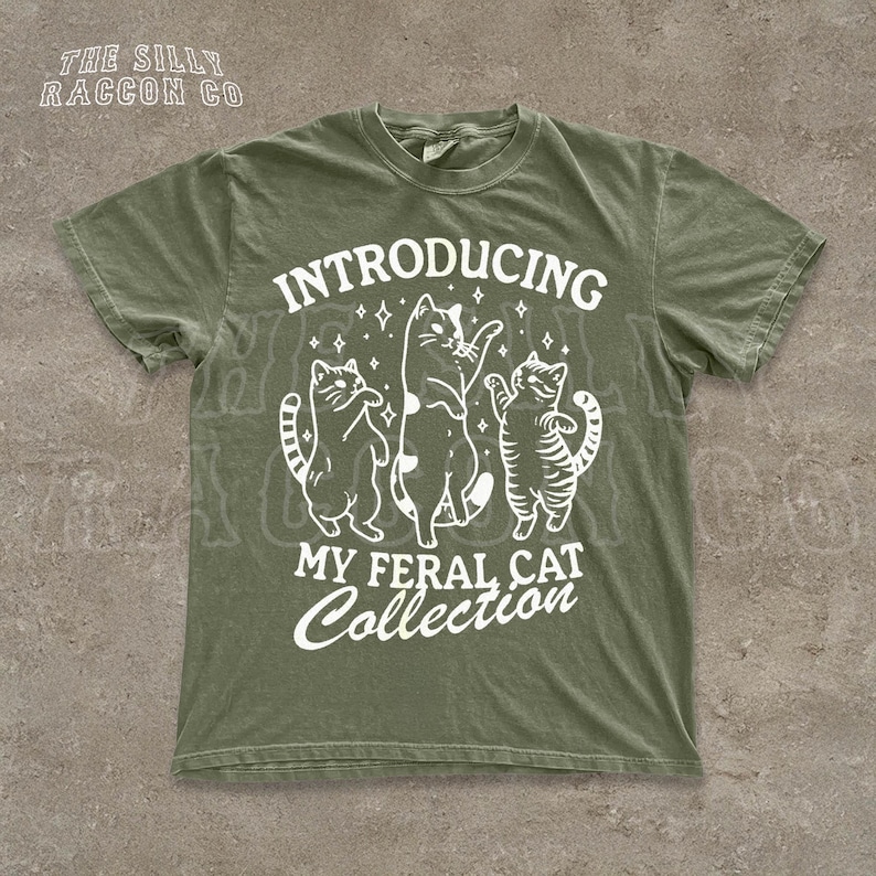 A green t-shirt with a graphic design featuring three cats and the text "INTRODUCING MY FERAL CAT COLLECTION" printed on it.