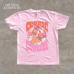 A pink t-shirt with a graphic design featuring a pair of cowboy boots and the text "COSMIC COWGIRL" printed on it.