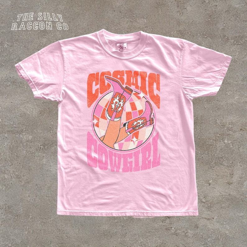 A pink t-shirt with a graphic design featuring a pair of cowboy boots and the text "COSMIC COWGIRL" printed on it.