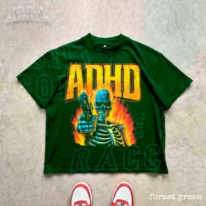 a green t-shirt with a graphic design depicting a skeleton holding a gun, with flames and text surrounding the image.