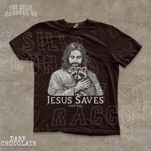 A t-shirt with a graphic design depicting a man holding a raccoon, along with the text "JESUS SAVES" and "LIKE YOURS".