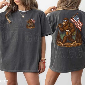 K&ouml;nnte beinhalten: Dunkelgraues T-Shirt mit einem Bigfoot-Motiv, das eine amerikanische Flagge und ein Bier h&auml;lt. Auf der Vorderseite des Shirts steht der Text "SALL ON CO". Auf der R&uuml;ckseite des Shirts befindet sich eine gr&ouml;&szlig;ere Bigfoot-Grafik mit der amerikanischen Flagge und Bier. Das Shirt hat eine lockere Passform.
