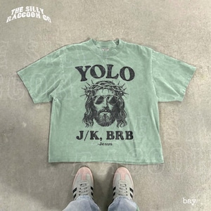 A green t-shirt with a graphic of Jesus wearing sunglasses and a crown, along with the text "YOLO J.K. BRB Jesus" printed on it. The t-shirt is laid out on a concrete floor, and a pair of sneakers is visible underneath it.