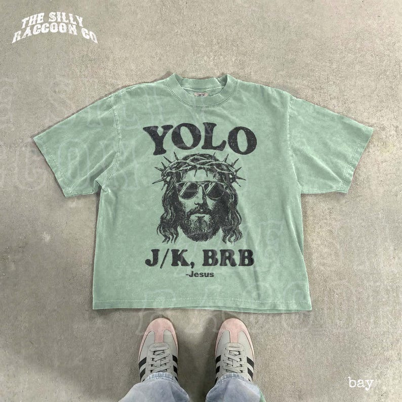 A green t-shirt with a graphic of Jesus wearing sunglasses and a crown, along with the text "YOLO J.K. BRB Jesus" printed on it. The t-shirt is laid out on a concrete floor, and a pair of sneakers is visible underneath it.