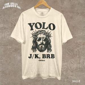 A white t-shirt with a graphic design featuring a stylized depiction of Jesus wearing sunglasses and a crown of thorns, along with the text "YOLO J/K, BRB - Jesus sand" printed on it.