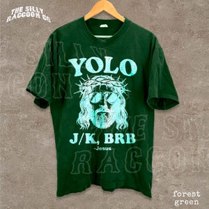 A green t-shirt with a graphic design featuring a stylized depiction of Jesus wearing sunglasses and a crown, along with the text "YOLO J/K, BRB Jesus" printed on it.