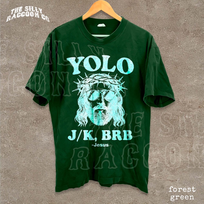 A green t-shirt with a graphic design featuring a stylized depiction of Jesus wearing sunglasses and a crown, along with the text "YOLO J/K, BRB Jesus" printed on it.