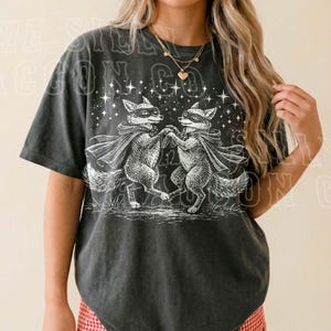A woman wearing a black t-shirt with a graphic design of two foxes dancing under a starry sky.
