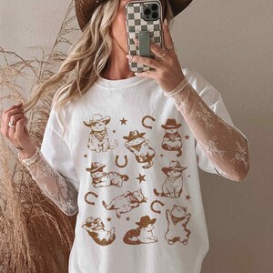 Vintage cowboy cat shirt for men and women featuring whimsical feline rodeo illustrations. Western-themed graphic t-shirt with cute kittens wearing cowboy hats, surrounded by horseshoes and stars. Soft, breathable fabric with a rustic, slightly distressed print for a retro aesthetic. Perfect for cat lovers, Western fashion enthusiasts, rodeo fans, and country music lovers. Unique gift idea for birthdays, holidays, or anyone who loves funny cat tees.