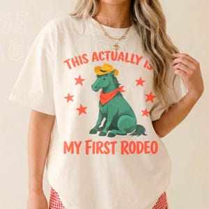 A woman wearing a white t-shirt with a graphic of a green horse and the text "This actually is my first rodeo" printed on it.